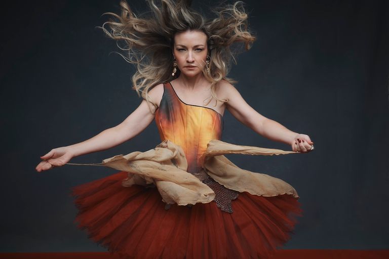 Australian singer Kate Miller-Heidke posing in an orange and red dress with windblown blonde hair against a dark backdrop.